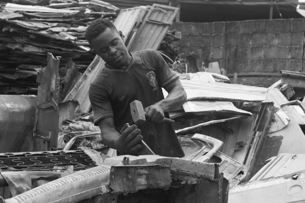 Black and white photograph of a man in a football jersey striking a chisel with a hammer as he breaks apart scrap metal, surrounded by towering piles of stripped car panels.