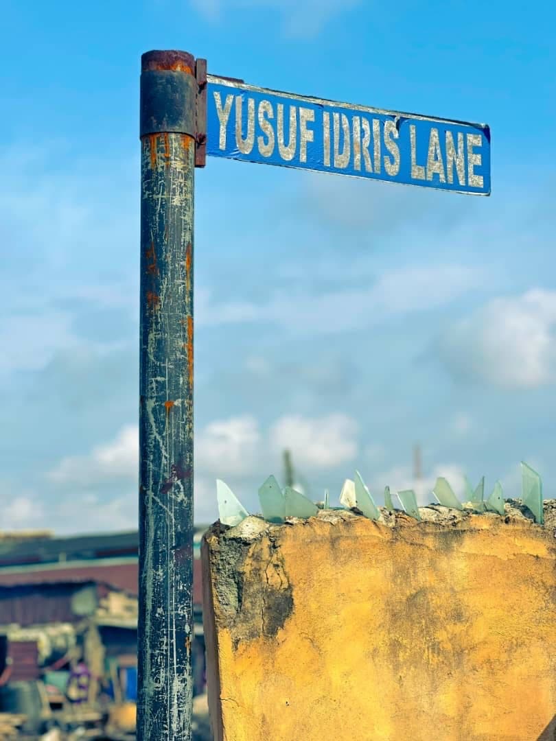 A weathered blue street sign reading “YUSUF IDRIS LANE” on a scratched metal pole against a bright, cloudy sky, above a wall topped with broken glass.