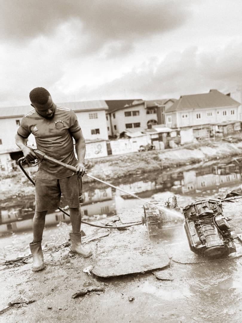 Sepia toned photograph of a man in a jersey, shorts and rubber boots using a pressure washer to clean a car engine on a muddy ledge, a densely built waterfront community behind him.