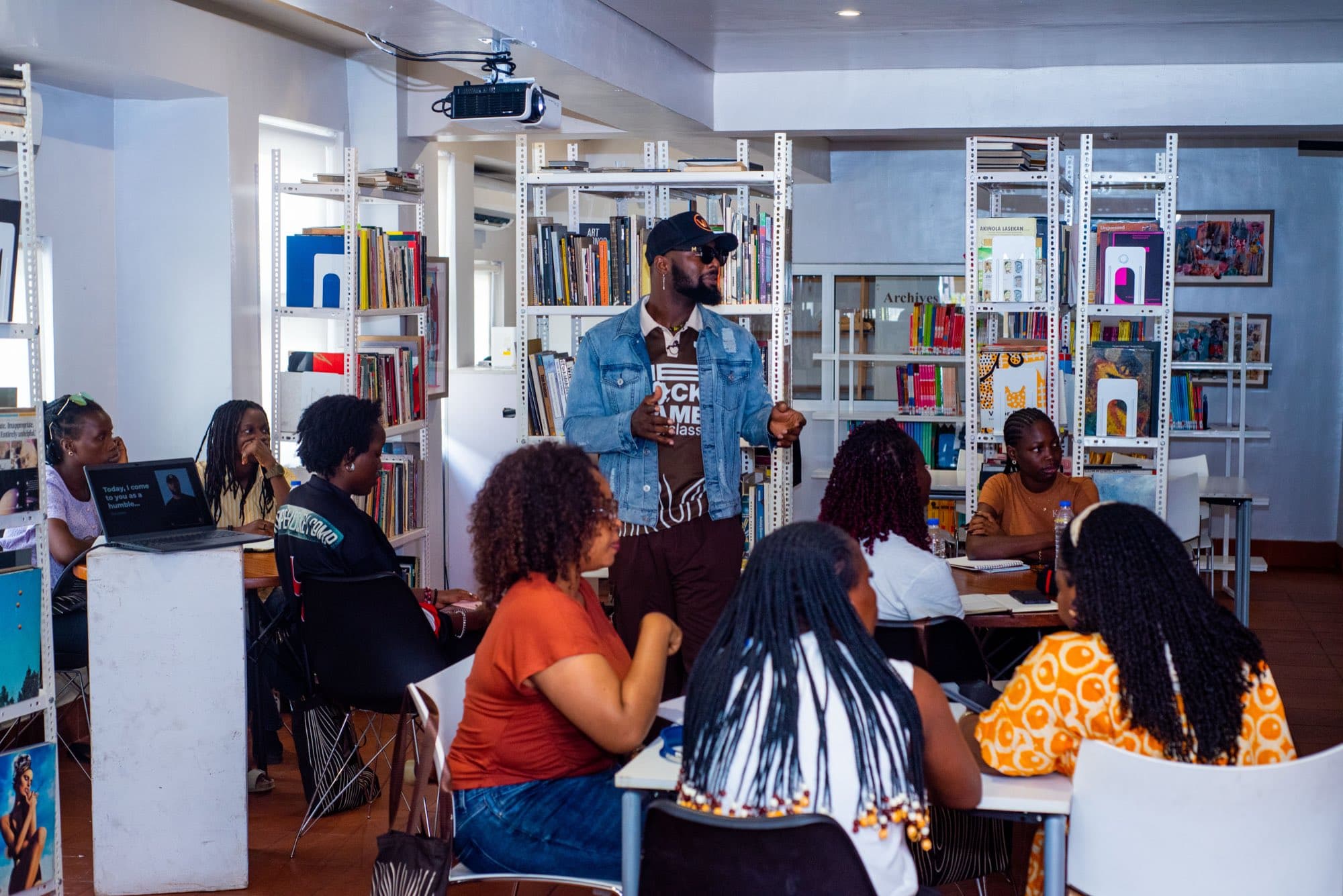 A guest speaker in a denim jacket and cap stands addressing seated participants in the book lined CCA Lagos library, a laptop slide reading “Today, I come to you as a humble…” beside him.