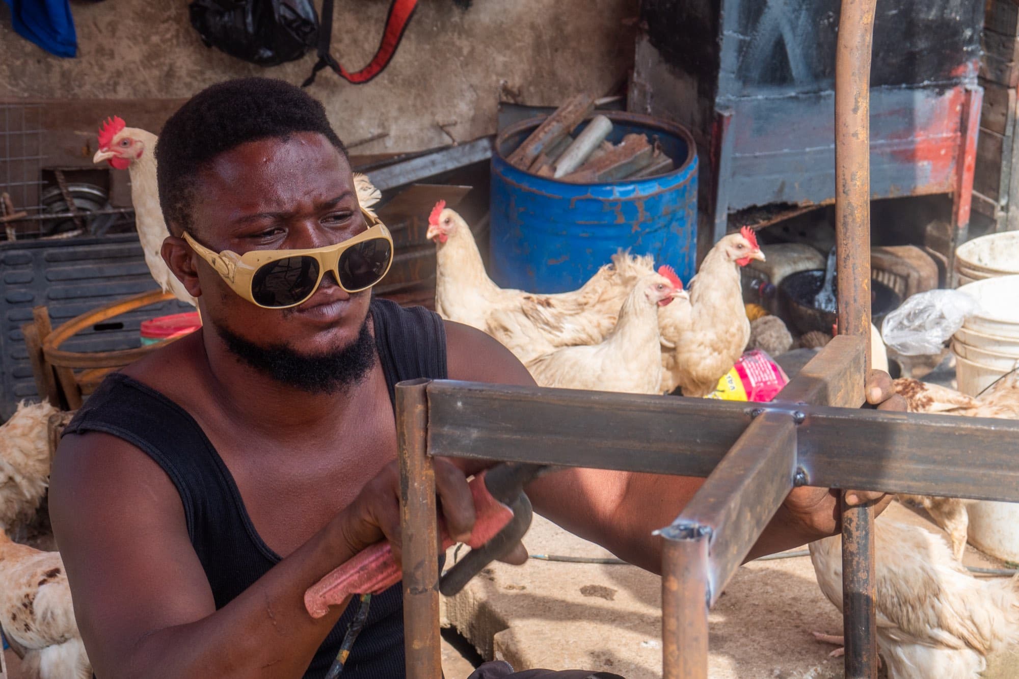 A young man welds a metal frame at a roadside workshop, wearing improvised sunglasses, with white chickens and a blue drum behind him.