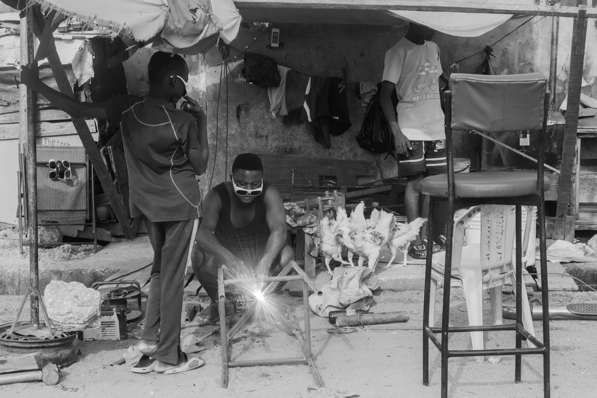 Black and white street scene in which a welder crouches over a bright arc weld while a boy looks on, with chickens and plastic stools around them.