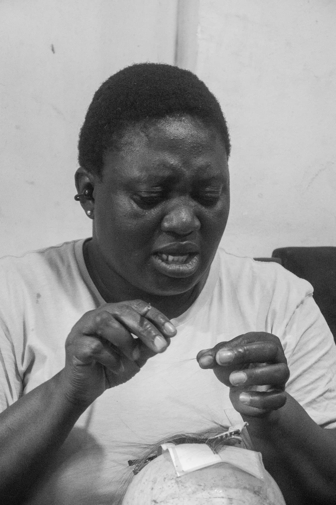 Black and white close portrait of a woman concentrating as she threads a needle, making a wig by hand.