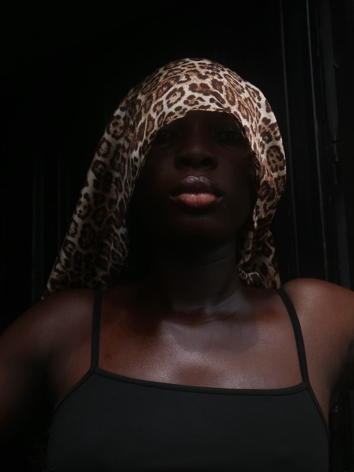 The woman wears the leopard print scarf wrapped over her head like a hood, her upper face in shadow and her gaze meeting the camera.