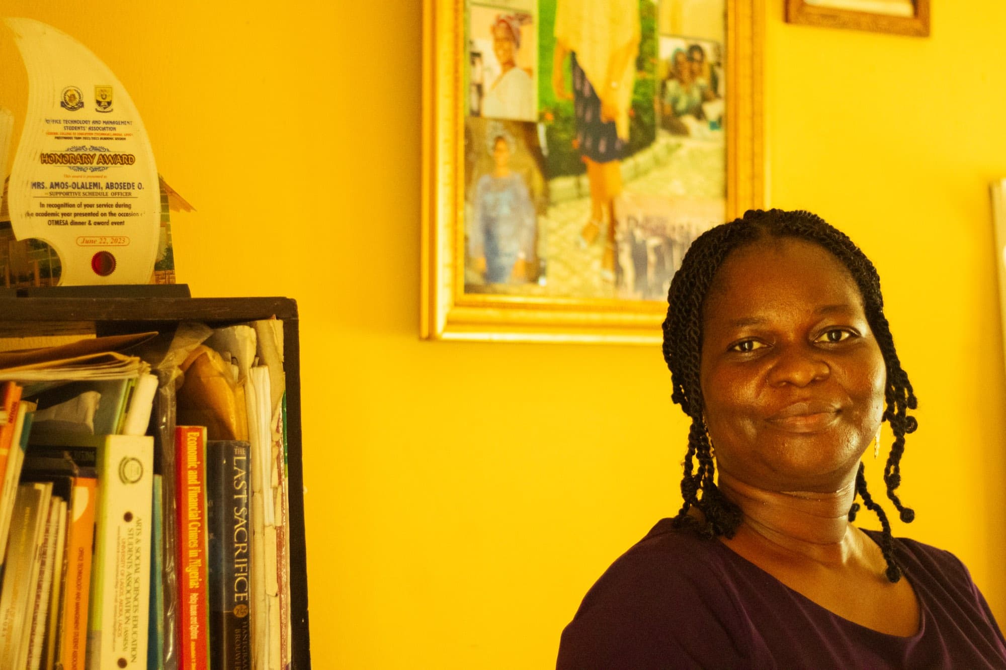 A woman with braided hair in a maroon top smiles beside a bookshelf holding an honorary award plaque and books, a framed photo collage on the yellow wall behind her.