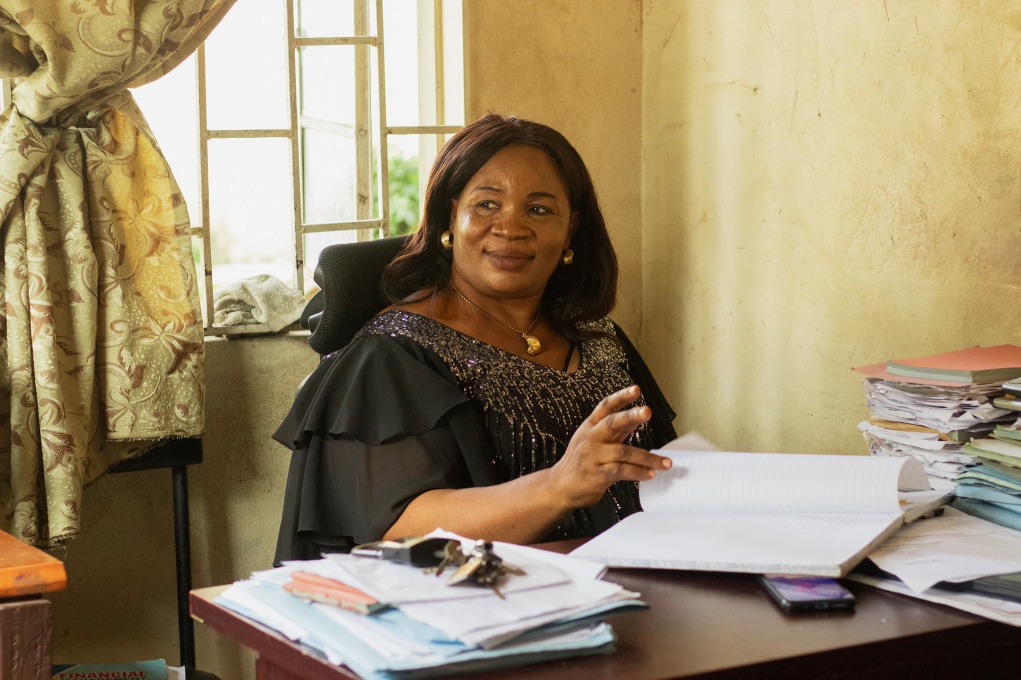 A woman in a black sequined dress sits at an office desk by a curtained window, an open ledger before her and tall stacks of files at her side.