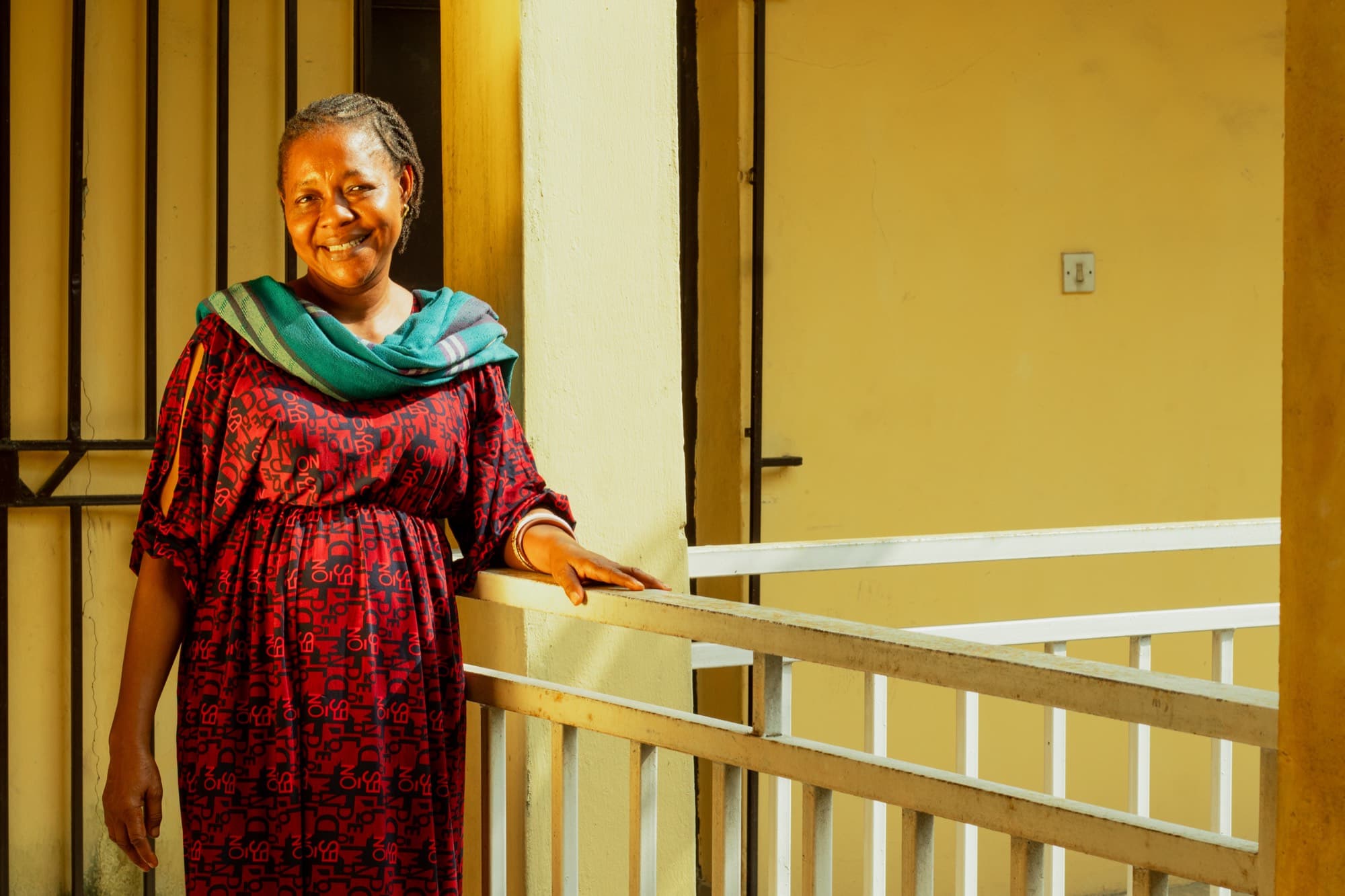 A smiling woman in a red and black patterned dress with a teal shawl stands at a white balcony railing along a yellow walled corridor.