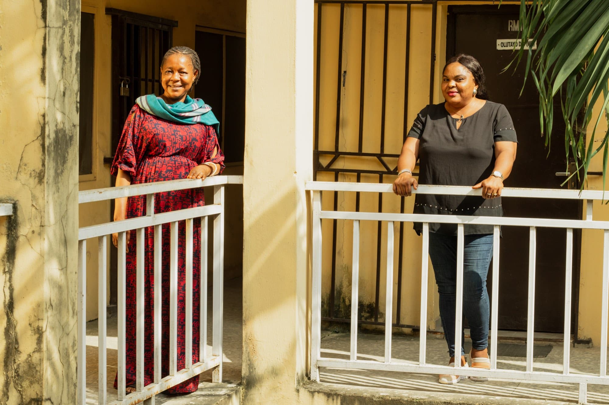 Two women stand at white balcony railings outside barred office doors in a sunlit yellow corridor; one in a red patterned dress and teal shawl, the other in a black top and jeans.
