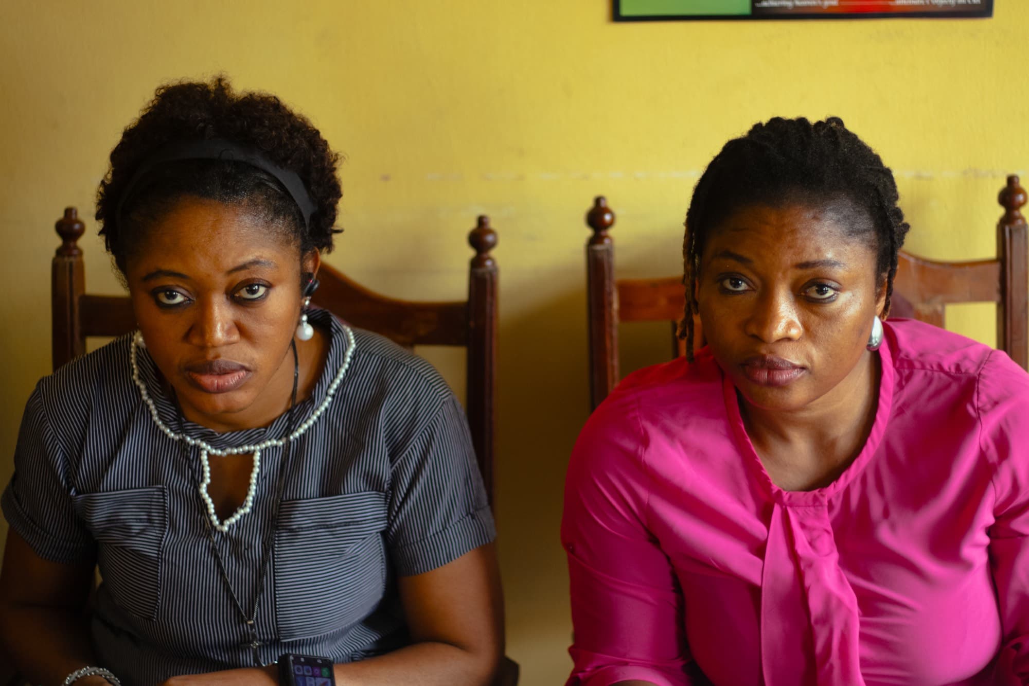 Two women sit on wooden chairs against a yellow wall, looking at the camera; one in a grey striped blouse with pearls, the other in a pink blouse.