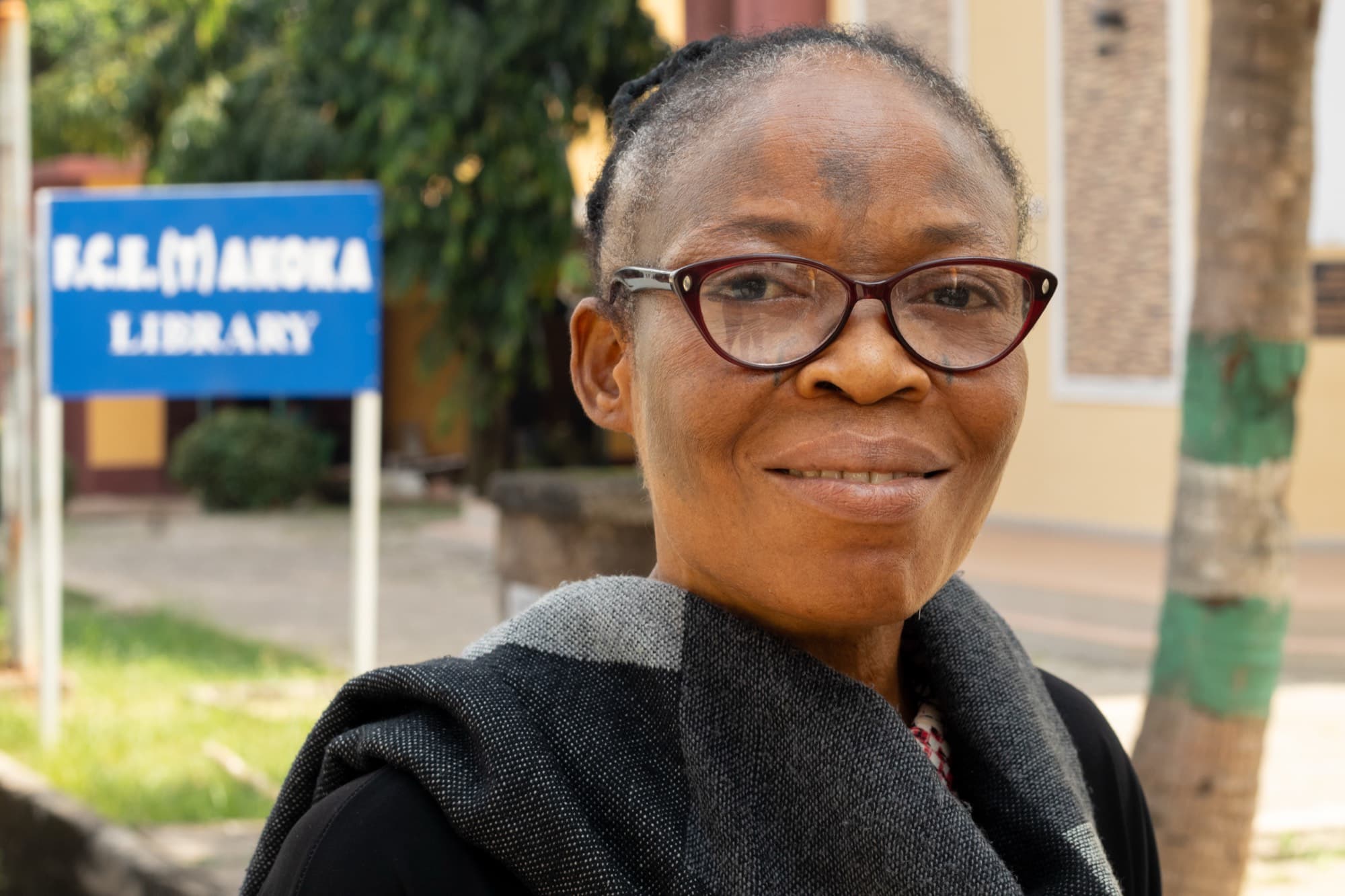 An older woman in red framed glasses and a grey and black shawl smiles outdoors in front of a blue sign reading “F.C.E.(T) Akoka Library.”
