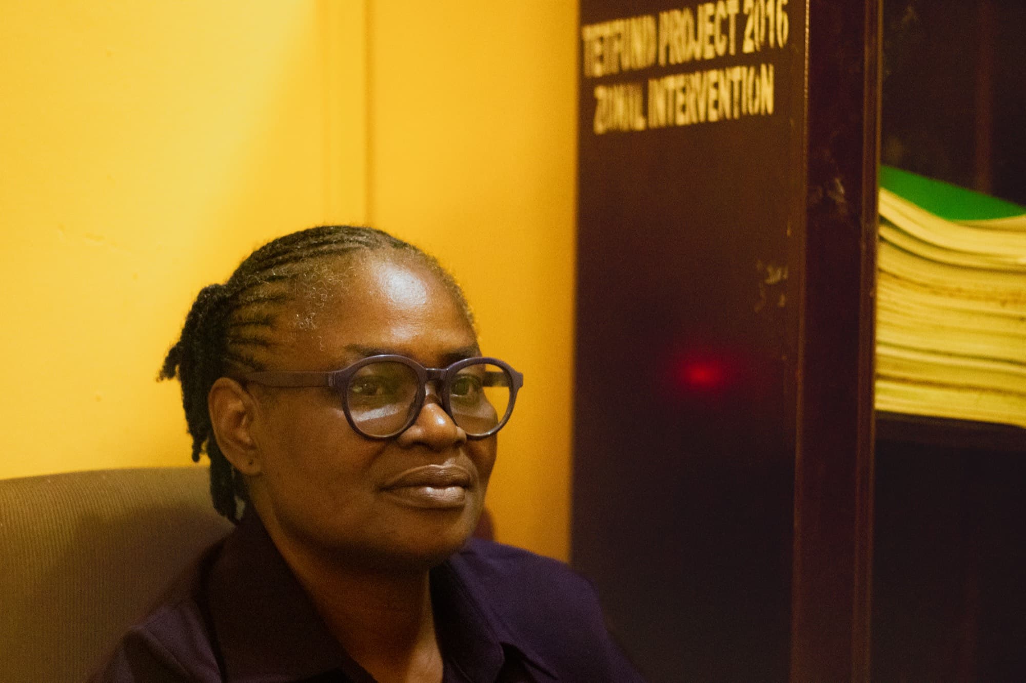 A woman with locs and round dark glasses in a dark shirt sits beside a wooden cabinet labelled “TETFund Project 2016” stacked with green files.