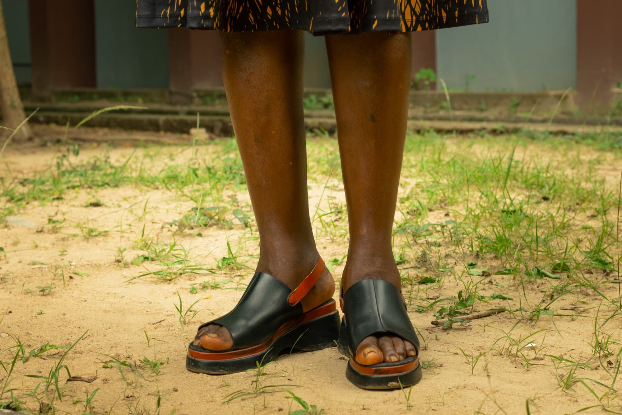 A close view of a woman's lower legs and feet in black and tan leather sandals, standing on sandy ground with sparse grass.