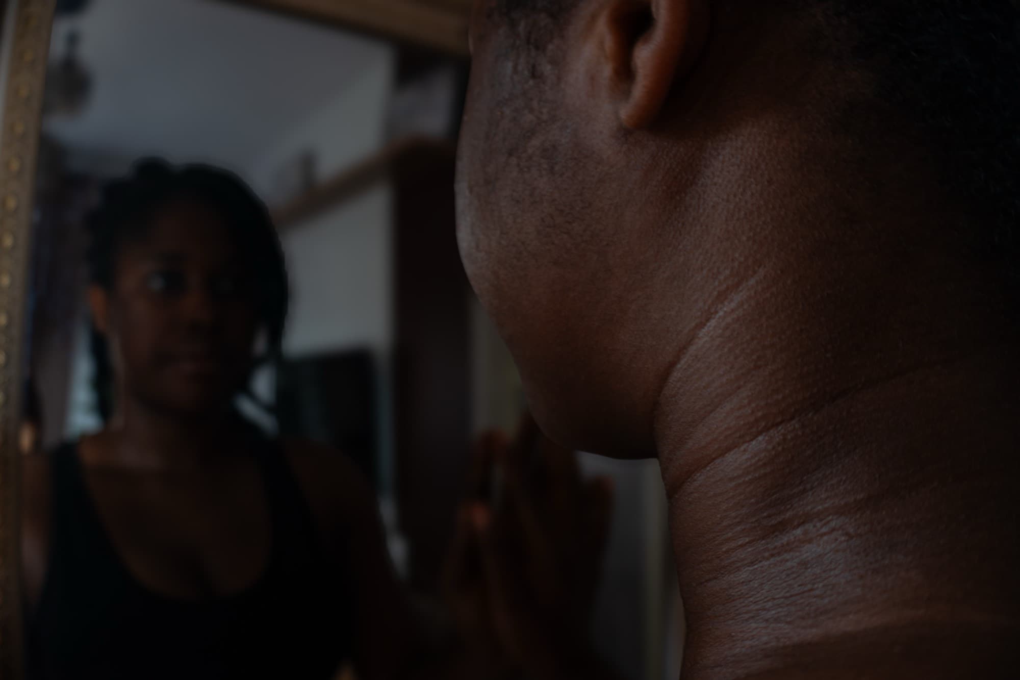 Extreme close up of the side of the woman's face and neck, with her uncertain reflection visible in a gold framed mirror behind her.