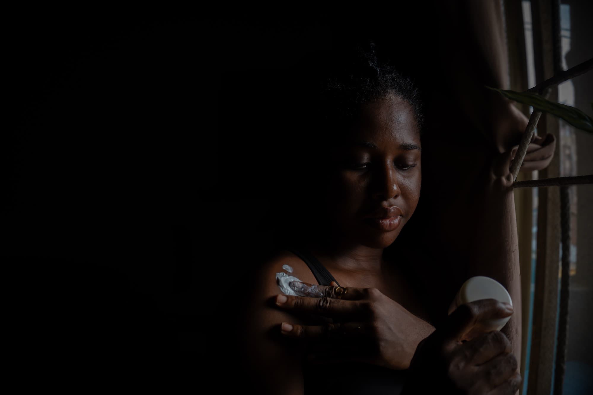 By a window in low light, the woman applies a white cream to her shoulder from a small tub, her eyes downcast.