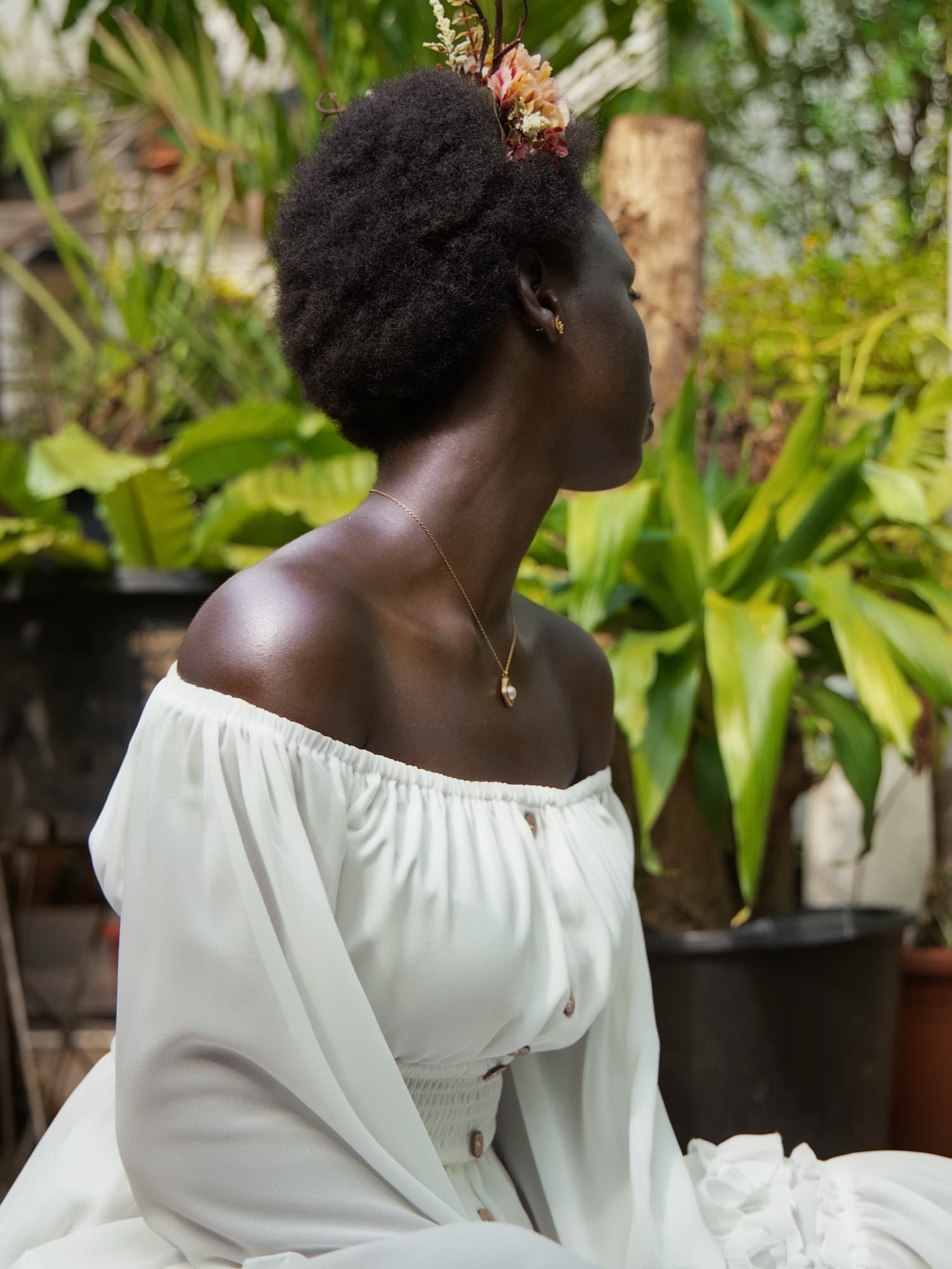 Profile view of the woman with peach and cream dried flowers tucked into her natural afro, wearing a white off shoulder blouse, against green tropical plants.