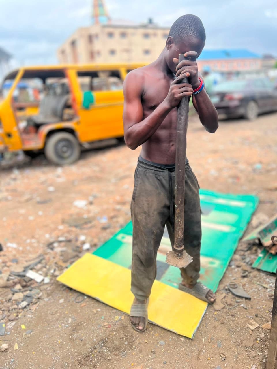 A shirtless young man rests his forehead on his hands atop a heavy metal breaker bar, standing on a green and yellow mat in a dusty roadside yard, with a yellow danfo bus and a car behind him.