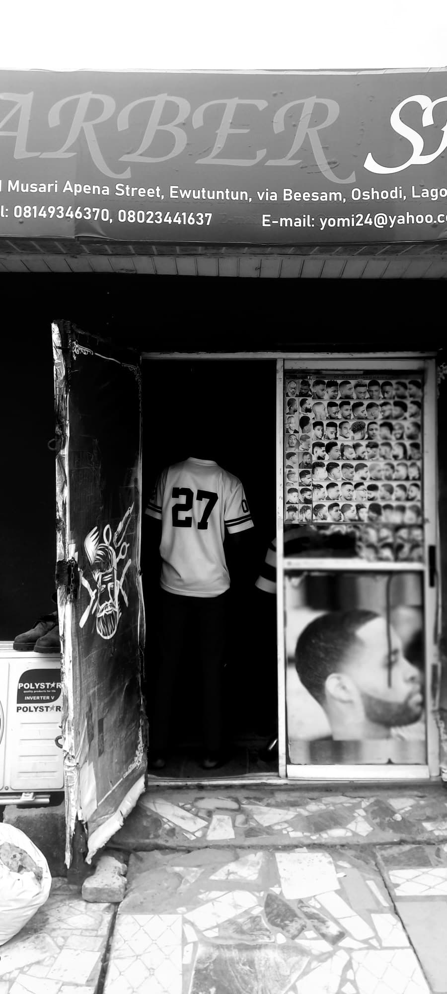 Black and white photograph of a Lagos barber shop storefront; a person in a jersey numbered 27 stands in the doorway beside a glass door showing a grid of haircut styles and a close up of a fresh fade.